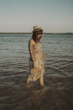 A young female with tattoos wearing a dress and straw hat on blurred ocean background