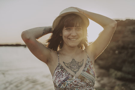 A young female with tattoos wearing a dress and straw hat on blurred ocean backgroundの写真素材