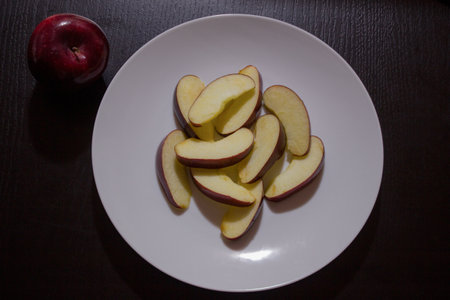 An overhead shot of a  dark red apple next to a plate of slices  on a dark table