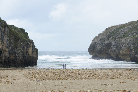 A couple standing on the coastline of the wavy sea between the two huge cliffsの写真素材