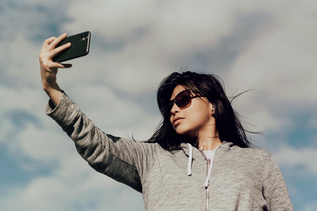 A young female wearing sunglasses taking a picture with her phone under the cloudy blue skyの写真素材
