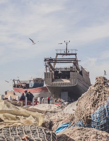 A vertical shot of an incomplete ship next to a lot of fishing nets with seagulls flying above