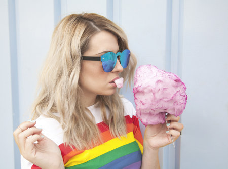 A portrait of a young caucasian blonde female with sunglasses eating cotton candy with the LGBT flag...