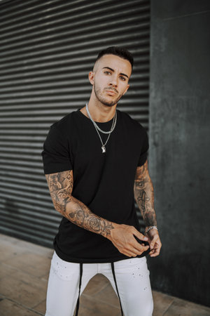A vertical shot of a young European tattooed hip male in a black shirt and white ripped jeans, and...