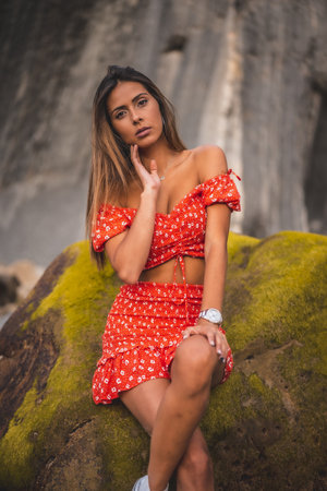 A shallow focus of a young female with long hair wearing a red dress and posing near the rocksの写真素材