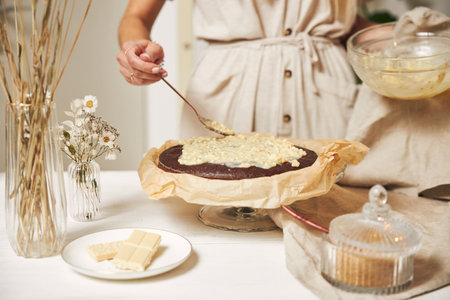 A young female baker making a delicious chocolate cake with cream on a white table
