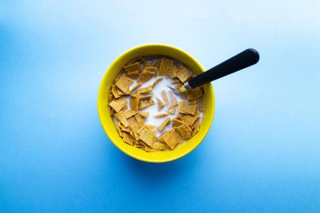 A top view of a bowl of cereal in square pieces with milk and spoon on a bowl isolated on a blue...