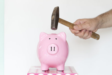 A caucasian male holding a hammer and about to break a piggy bank, in front of a white background