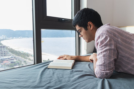 An Indian male reading a book on the bed by a window with a nice viewの写真素材