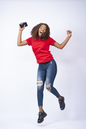 A vertical shot of a happy young black lady jumping in excitement and holding a phone
