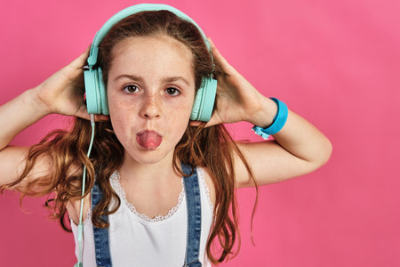 A little girl posing with headphones with her tongue out on a pink background
