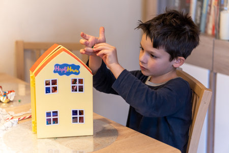 POZNAN, POLAND - Dec 25, 2020: Polish young Caucasian boy playing with a plastic Peppa Pig toy house by a wooden table.の写真素材
