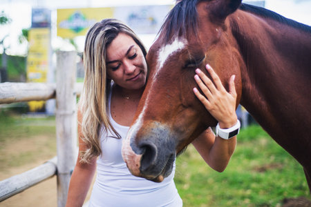 A shallow focus of a young Hispanic female petting a brown horse in a farm under the sunlightの写真素材
