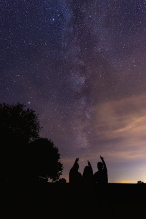 A vertical shot of silhouettes of three people pointing at the milky way in the night skyの写真素材