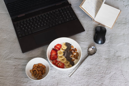 A high angle shot of a bowl of yogurt with fruits and nuts on the table with a laptop on itの写真素材