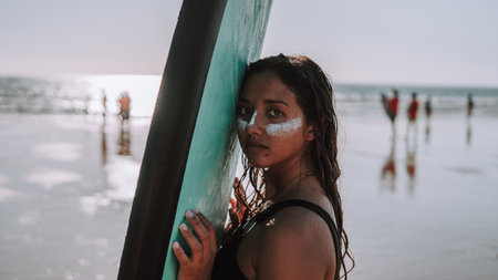 A European surfer female with a sunscreen on her face holding her surfboard with the ocean in the backgroundの写真素材