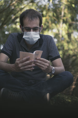 A man sitting on a rock reading a booklet and wearing a facemask on a blurred backgroundの写真素材