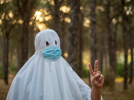 A shallow focus shot of a man dressed like a bedsheet ghost, also wearing a facemask and holding a peace signの写真素材