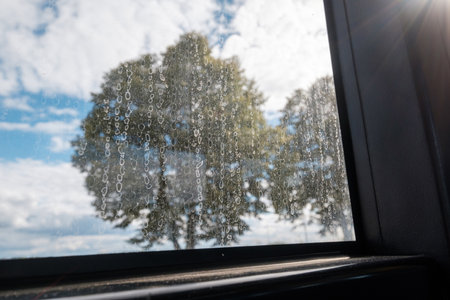 A closeup shot of a window with stains from dried water and seen green trees and cloudy sky