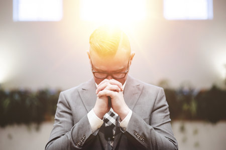 A young male in a formal outfit praying at the church with the bright sun in the backgroundの写真素材