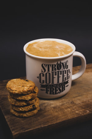 A vertical shot of a cup of coffee with some cookies on a wooden tray in a dark studio