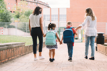 Two women and two children holding hands from back go to school for first time after coronavirus pandemic. Mothers accompanies the students along way.の写真素材