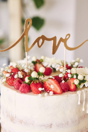 A selective focus shot of delicious white wedding cake with red berries, flowers and cake topperの写真素材