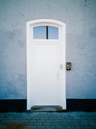 A vertical shot of a white door on a grey wall with a doorbell on the wall