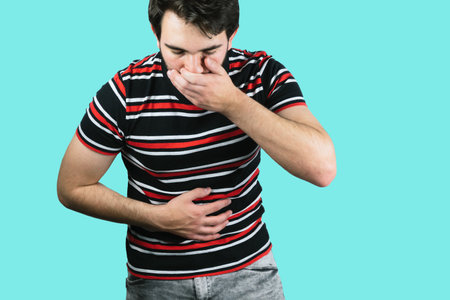 A young male holding his belly with one hand and the mouth with the other throwing up from food poisoning with a blue backgroundの写真素材