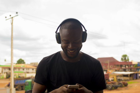 A young African male standing outside, using his wireless headphone to listen to music on the phoneの写真素材