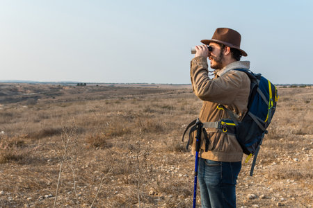 A Caucasian hiker man with a beard, wearing a brown jacket, backpack, looking through binocularsの写真素材