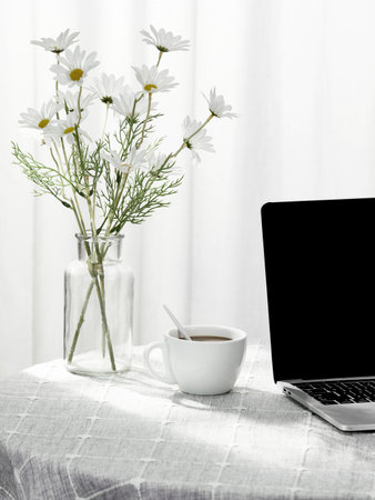 A vertical shot of a laptop and a cup of coffee next to a vase full of flowers, indoors
