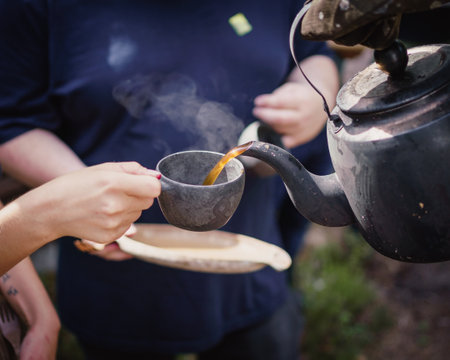 A closeup of a person pouring hot tea from a pot into a ceramic cup under the sunlight