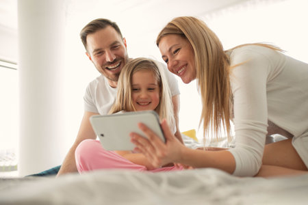 A happy young caucasian parents laughing while watching the tablet screen with their little...