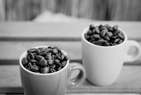 A grayscale closeup shot of two full cups with roasted coffee beans on a wooden tableの写真素材