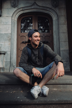 A vertical shot of a young South American man sitting against an old door with a backpackの写真素材