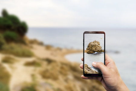 A closeup of a hand holding a phone taking a photo of a beachの写真素材