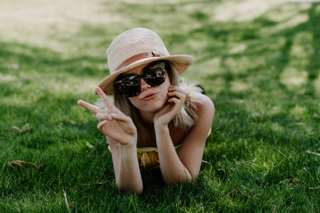 A closeup shot of a beautiful young lady wearing a beach hat and sunglasses lying on the grassの写真素材