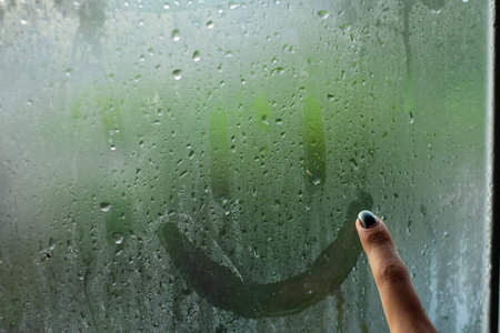 A female finger drawing a smiley on wet window with rain drops on the other side - concept of sadnessの写真素材