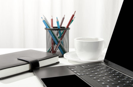 A closeup of a laptop, a cup of coffee, a notebook and pencil holder on a white desk, indoorsの写真素材