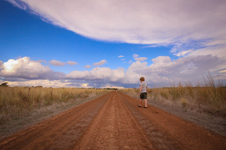 A little boy walking through a road in a rural field under the cloudy skyの写真素材