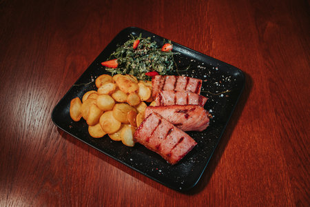 A top view closeup of potatoes with grilled fish fillet and herbs on a black plate on a wooden tableの写真素材