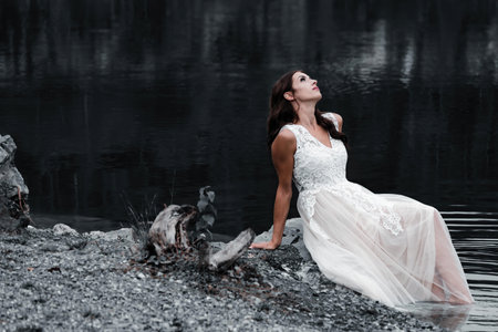 A beautiful shot of a young woman in a white dress sitting by the lakeの写真素材