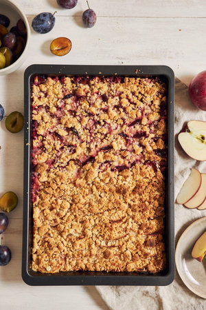 A top view of freshly baked plum and apple cake in a pan and fresh plums and apples on a white tableの写真素材