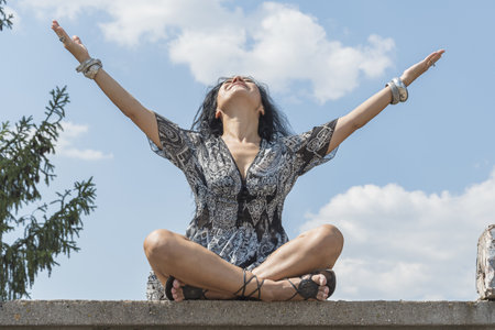 A young brunette girl sits in a lotus position on a concrete surface and raises her arms and head up to the sky. Yoga class.の写真素材