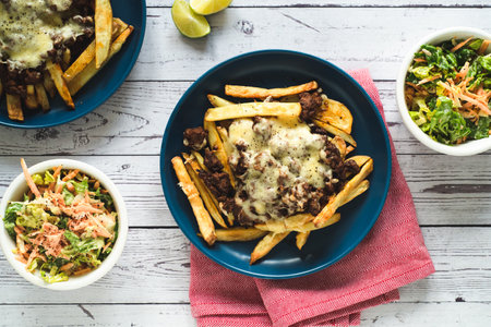 A high angle shot of servings of french fries with fried meat and cheese, salads in bowls, and limes