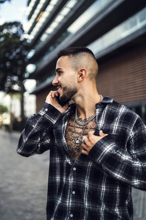 A vertical closeup shot of a young handsome European man with tattoos in a plaid patterned shirt talking on the phone in the streetの写真素材