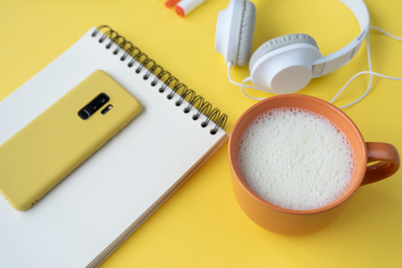 A high angle shot of a smartphone, a journal, headphones, pens, and a cup of milk on a yellow table