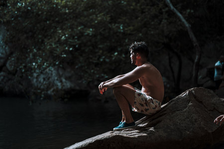 A young attractive man in a swimsuit sitting on a rock in a forest lake and enjoying the nature sceneの写真素材