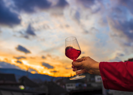 A closeup shot of a person's hand holding a glass of red wine with the beautiful sunset in the backgroundの写真素材